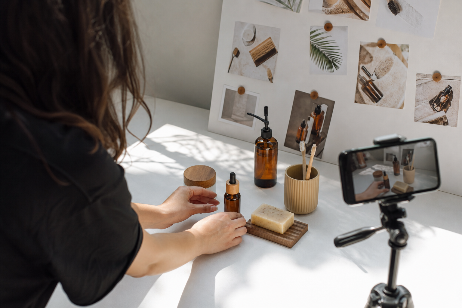 Flat-lay of a product photography shoot in progress, with a smartphone on a tripod, ecommerce products arranged on a white surface, and a mood board pinned nearby under natural window light